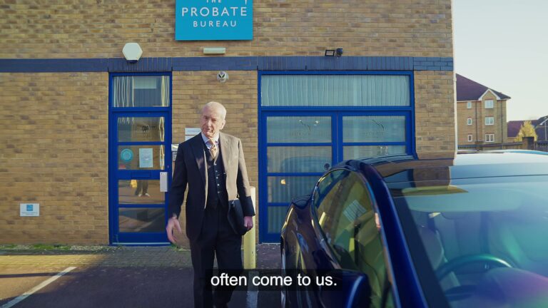 Older man in a dark suit stands outside a brick building with blue-framed doors labeled The Probate Bureau, near a parked car. – Lumira Studio